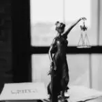 Grayscale photo of Lady Justice statue beside legal documents on a wooden table.
