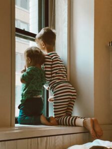 Two kids in pajamas curiously peering out of a window inside their home.